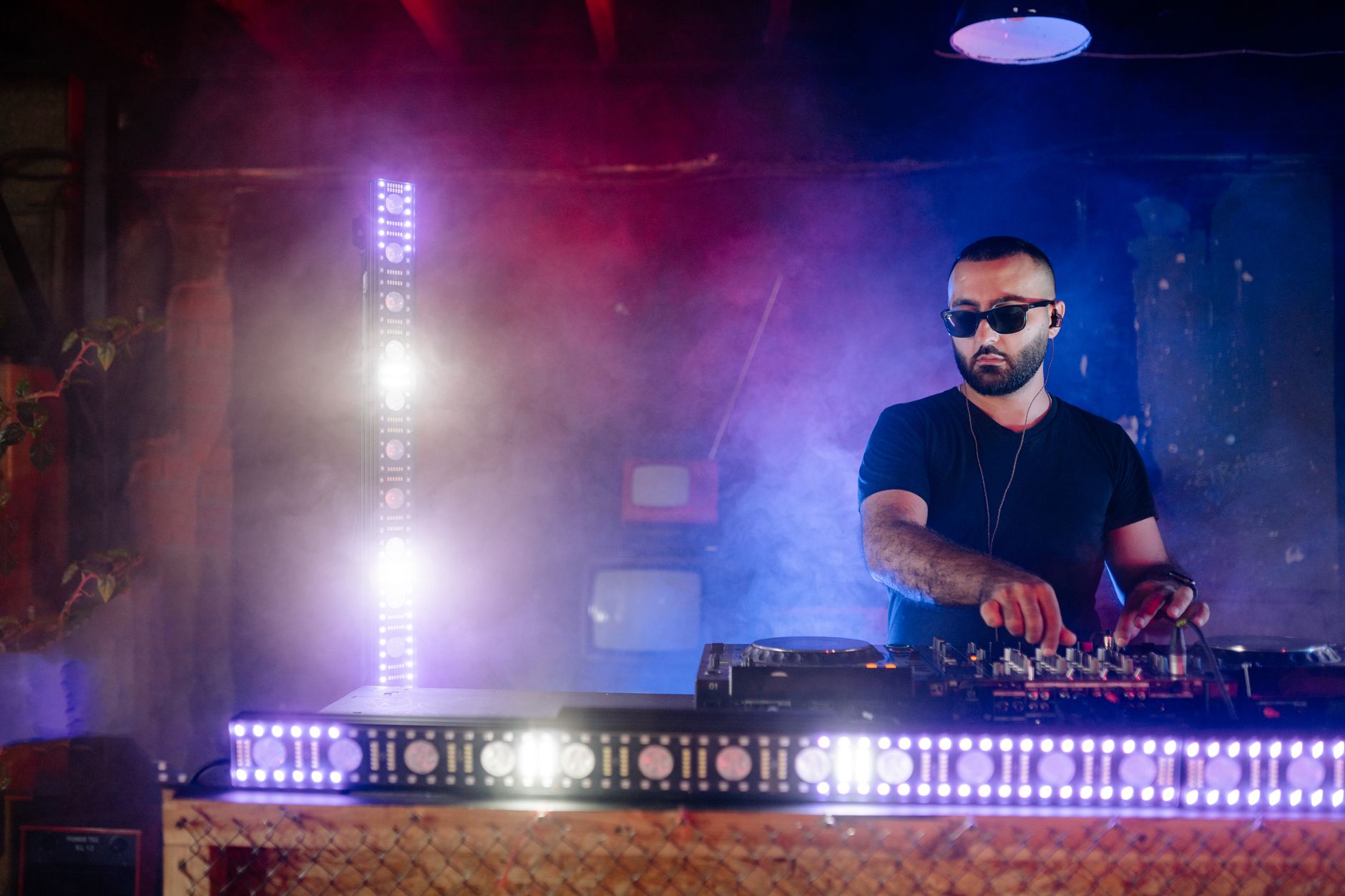 Un DJ portant des lunettes de soleil mixe de la musique sur une platine, dans une ambiance de boîte de nuit avec des lumières vives et de la fumée.